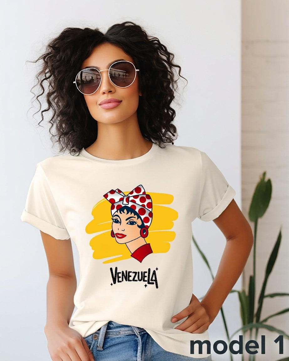 A woman wearing a cream-colored graphic t-shirt with a stylized illustration of a Venezuelan woman in a red and white polka-dot headscarf and the word 'VENEZUELA' printed below. She is wearing round sunglasses and blue jeans, standing in a brightly lit indoor setting.