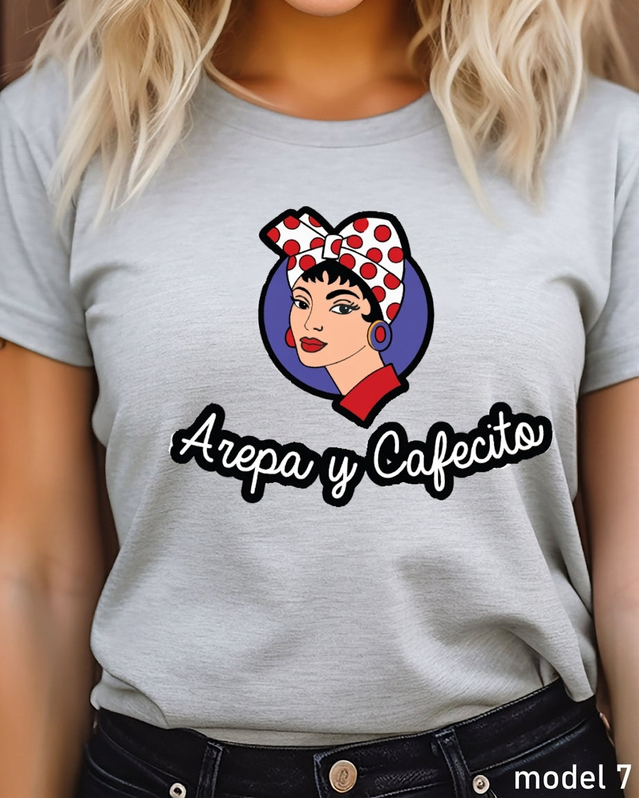 A woman wearing a light gray heather t-shirt with a graphic of a woman in a red and white polka dot headscarf and the text 'Arepa y Cafecito'.